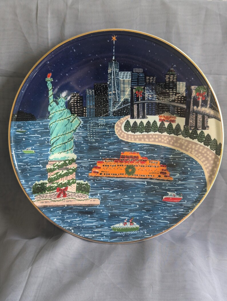 May include: A decorative plate featuring a winter scene of New York City. The plate depicts the Statue of Liberty, the city skyline, and an orange ferry boat on a blue background. The plate has a gold rim.