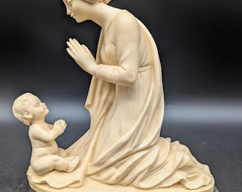 May include: A cream-colored statue depicting a woman kneeling with clasped hands, looking down at a baby also with clasped hands. The figures are on a round base atop a rectangular, dark gray marble base. The woman wears a draped gown.