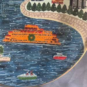 May include: A decorative plate featuring a winter scene with an orange ferry boat on blue water. The plate has a gold rim and depicts a city skyline, a pathway, and small boats. A green wreath adorns the ferry.