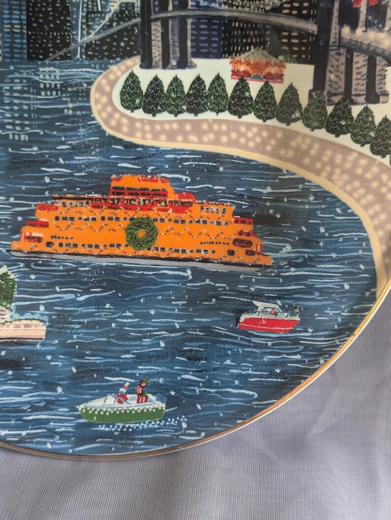 May include: A decorative plate featuring a winter scene with an orange ferry boat on blue water. The plate has a gold rim and depicts a city skyline, a pathway, and small boats. A green wreath adorns the ferry.