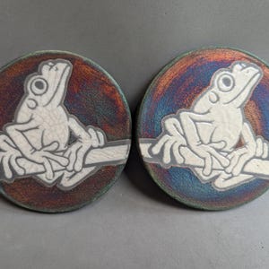 May include: Two circular ear plugs featuring a white, crackled-texture frog illustration perched on a branch. The background of each plug displays a metallic, iridescent finish with shifting hues of deep blue, purple, and copper tones.