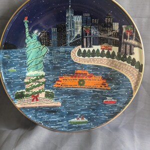 May include: Decorative plate featuring a winter scene of New York City. The Statue of Liberty is adorned with garland. The plate has a gold rim and depicts the city skyline, a ferry, and boats on the water.
