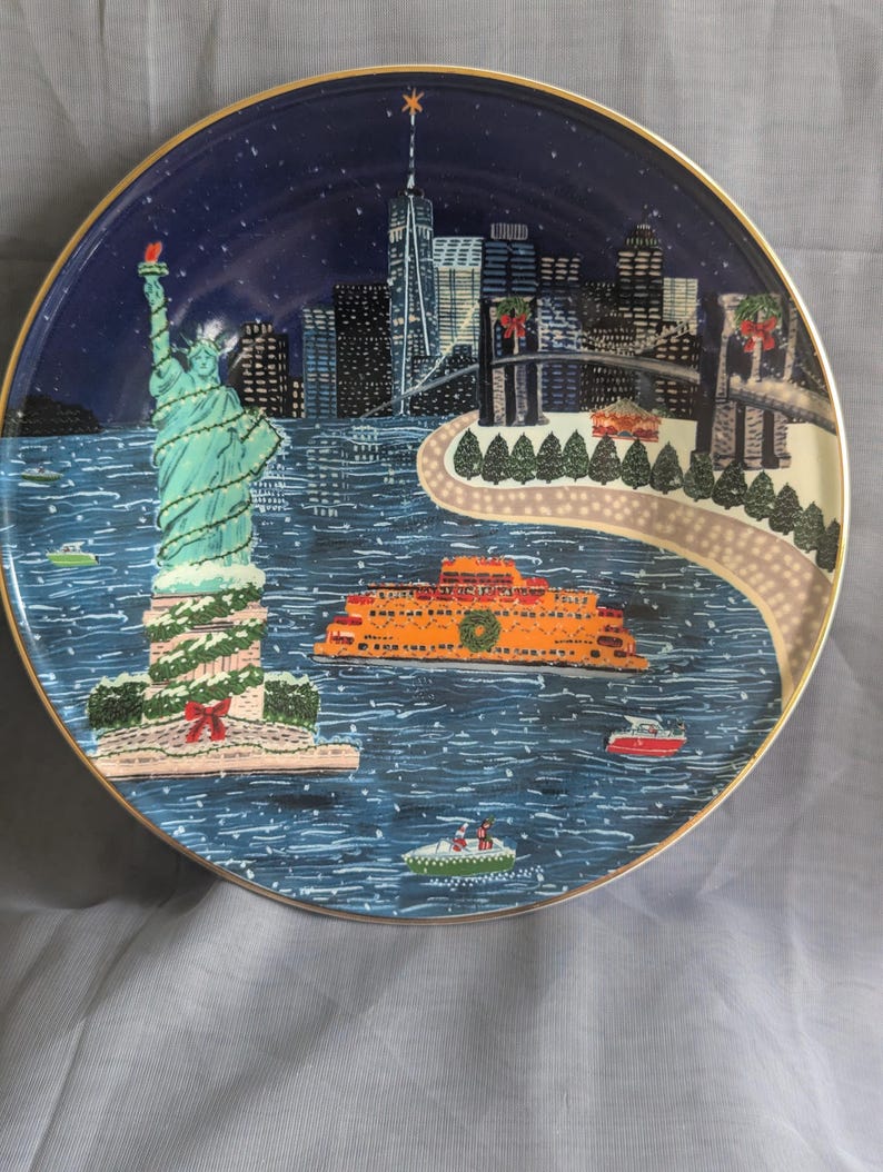 May include: Decorative plate featuring a winter scene of New York City. The Statue of Liberty is adorned with garland. The plate has a gold rim and depicts the city skyline, a ferry, and boats on the water.