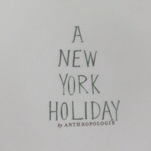 May include: A white ceramic plate with the text "A NEW YORK HOLIDAY" in dark green lettering. The words "by ANTHROPOLOGIE" are below the main text.