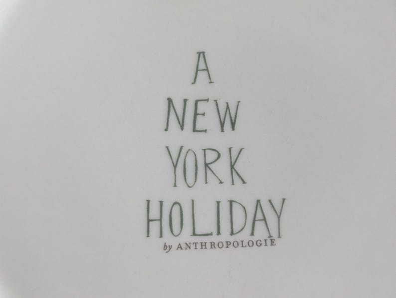 May include: A white ceramic plate with the text "A NEW YORK HOLIDAY" in dark green lettering. The words "by ANTHROPOLOGIE" are below the main text.