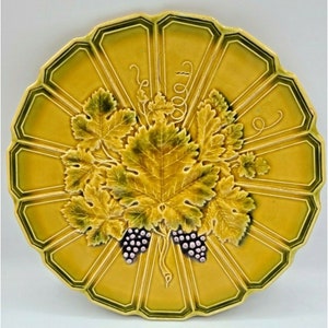 May include: A decorative ceramic plate with a yellow and green color scheme. The plate features a raised relief design of grape leaves and bunches of grapes. The plate has a scalloped edge.