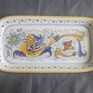 May include: A rectangular ceramic serving tray with handles, featuring a hand-painted design of a yellow and blue mythical sea creature, a teal flower, and scrolling flourishes. The border is yellow with a thin black line and orange dots.