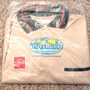 May include: A tan polo shirt in a plastic bag. The shirt has a green and red collar, a pocket, and the "Riverside" logo with a world map. The Coca-Cola logo is also visible.