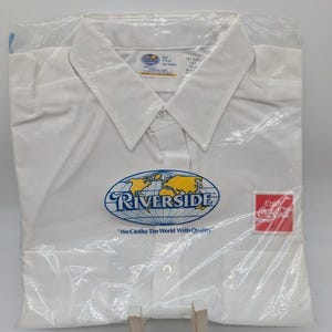 May include: White collared shirt in a plastic bag, featuring the "Riverside" logo with a world map design in blue and yellow, and the Coca-Cola logo. The shirt has a button-down front and the text "We Clothe The World With Quality".