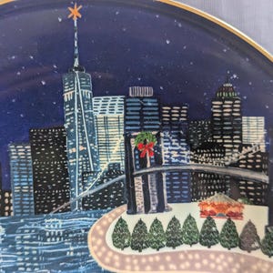 May include: Decorative plate featuring a nighttime cityscape with the New York City skyline. The plate has a blue background with a gold rim and a star on top of the tallest building. The image includes the Brooklyn Bridge and Christmas decorations.