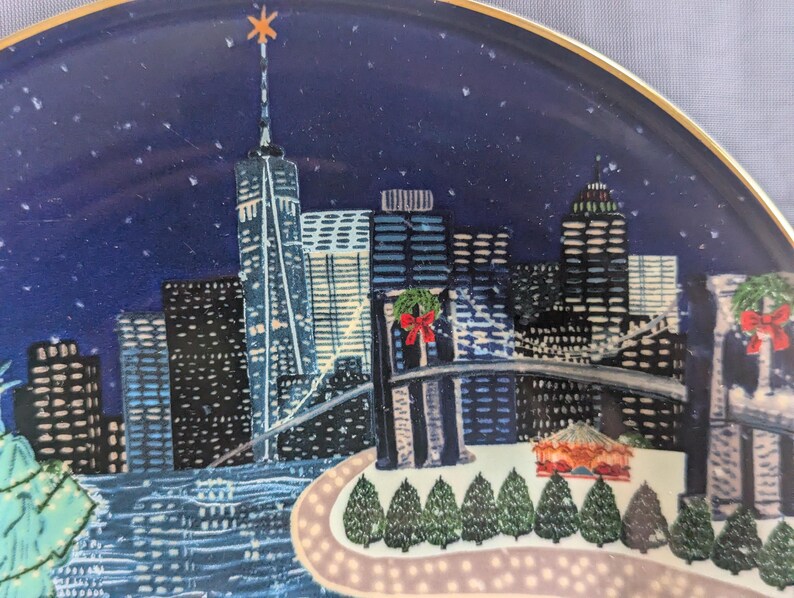 May include: Decorative plate featuring a nighttime cityscape with the New York City skyline. The plate has a blue background with a gold rim and a star on top of the tallest building. The image includes the Brooklyn Bridge and Christmas decorations.