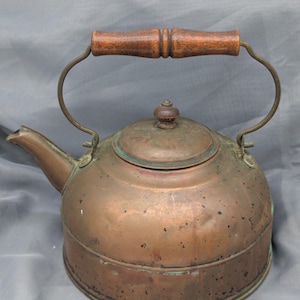 May include: A vintage copper kettle with a wooden handle and a curved metal handle. The kettle has a round body, a spout, and a lid with a small wooden knob. The copper has a patina, showing signs of age and use.