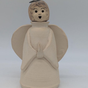 May include: A ceramic angel figurine with a surprised expression. The angel has a light beige body, wings, and a head with detailed facial features. A metal halo and a wick are attached to the head. The angel's hands are clasped in front.
