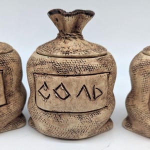 May include: Three ceramic salt and pepper shakers shaped like burlap sacks with lids. The shakers are labeled "перец" (pepper), "соль" (salt), and "горчица" (mustard).