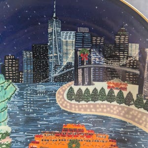May include: Decorative plate featuring a winter scene of New York City. The plate depicts the city skyline, a ferry, and the Statue of Liberty, all under a starry night sky. The plate has a gold rim.