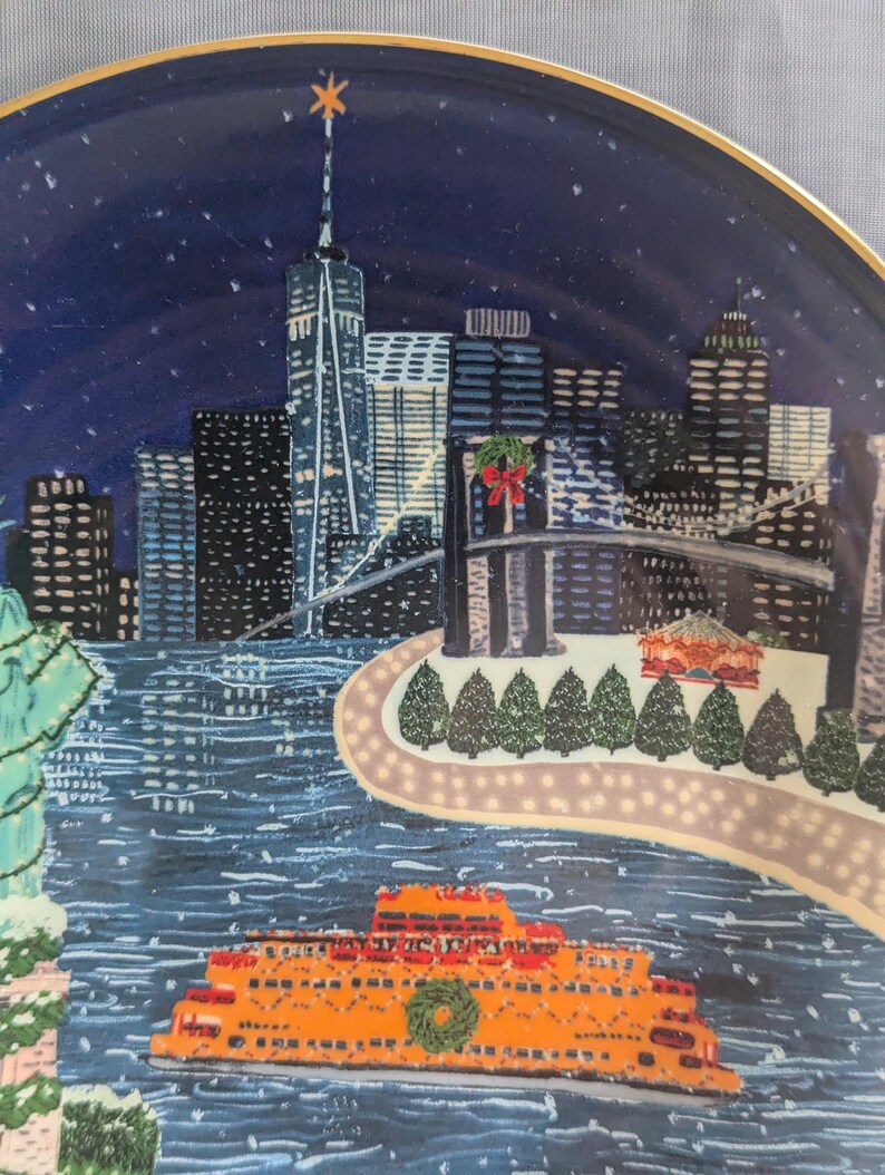 May include: Decorative plate featuring a winter scene of New York City. The plate depicts the city skyline, a ferry, and the Statue of Liberty, all under a starry night sky. The plate has a gold rim.