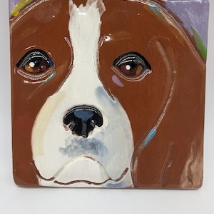 May include: Square ceramic tile featuring a dog's face. The dog has brown fur with white markings on its face and a black nose. The eyes are detailed with brown and black, and the tile has a glossy finish. The background has a purple and green color.