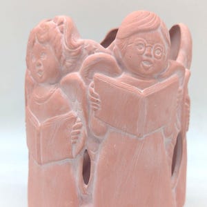 May include: A pink ceramic candle holder featuring a choir of three angel figures, each holding a book. The figures are sculpted with detailed facial features and wings, and the holder has openings for light to shine through.
