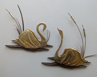 May include: Two wall-mounted decorative pieces featuring gold-toned metal swans with textured feathers. Each swan is positioned on a dark brown wooden base shaped like water, accompanied by tall, thin metal cattail stalks with gold-colored tips.