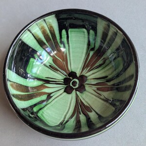 May include: A round ceramic bowl featuring a glossy finish with a radiating pattern of light green, dark brown, and black streaks. The interior base of the bowl is decorated with a small, dark brown five-petaled flower design at the center.