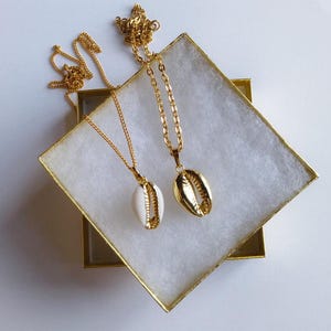 May include: Two gold-colored necklaces with seashell pendants rest inside a white box with a gold trim. One pendant is white with gold trim, and the other is solid gold. The necklaces have delicate gold chains.