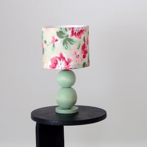 May include: A miniature lamp with a floral lampshade featuring pink roses and green leaves. The lamp has a light green base and sits on a small black table. The lamp is a decorative item.