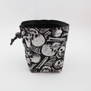 Pode incluir: Saco de cordão preto com um padrão de caveiras e ossos brancos. O saco tem um fecho de cordão preto.
