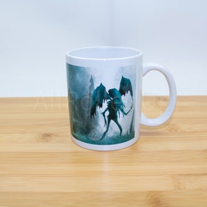 Può includere: Tazza in ceramica bianca con una grafica di una creatura alata in una foresta oscura e nebbiosa. La creatura è blu e verde e ha un corpo lungo e sottile.