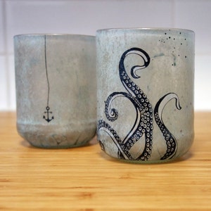 May include: Two glass cups with a light blue glaze. One cup has a black anchor design, the other has a black octopus design.