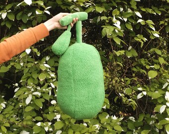 May include: A green plush toy shaped like a pear with a stem and a small leaf. The toy is being held by a hand in front of a green leafy background.