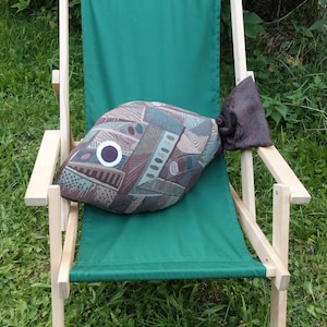 May include: A brown and green patterned fish-shaped pillow sits on a green canvas folding chair. The chair is made of light wood and is sitting on a bed of green grass.