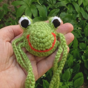 May include: Green crocheted frog with a red smiling mouth and white eyes. The frog has long green crocheted legs.