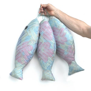 Peut inclure: Trois coussins en forme de poisson &agrave; motifs bleus et roses sont suspendus &agrave; une ficelle tenue par une main. Les coussins sont faits d'un tissu doux et textur&eacute;.