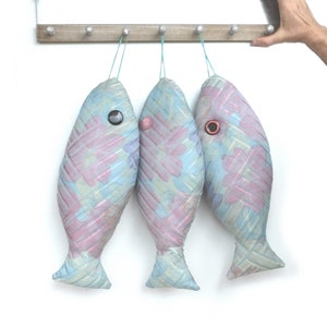 Peut inclure: Trois ornements en forme de poisson de couleur bleu pastel et rose suspendus &agrave; un support en bois. Chaque poisson a un &oelig;il en bouton et est fait d'un tissu tiss&eacute;.