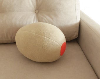 May include: A beige plush olive-shaped pillow with a red circle on one side, resting on a beige sofa.