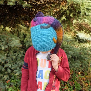 May include: A blue, purple, and orange crocheted hat shaped like a lizard or dragon. The hat covers the head and face, with the tail extending down the side. The hat is worn over a red jacket and a white t-shirt with colorful text.