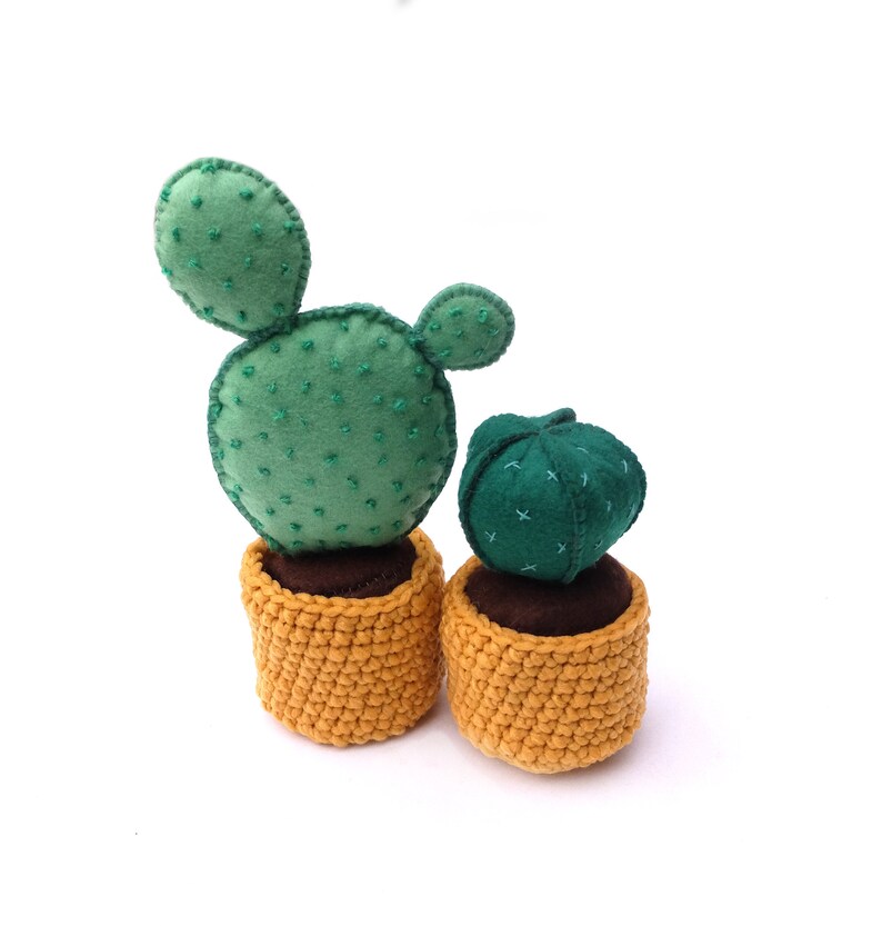 May include: Two felt cactus plants in crocheted pots. The larger cactus is green with a single arm and the smaller cactus is a round, dark green ball. Both pots are a mustard yellow colour.