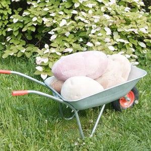 May include: A gray wheelbarrow filled with three large, soft, plush pillows. The pillows are pink, beige, and white. The wheelbarrow is sitting on a grassy lawn.