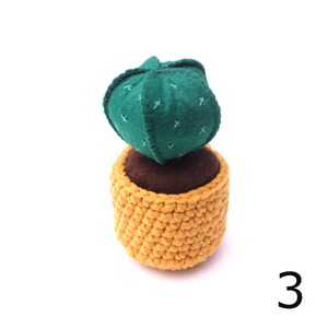May include: A green felt cactus with brown felt soil in a yellow crocheted pot. The cactus has a stitched pattern of crosses.