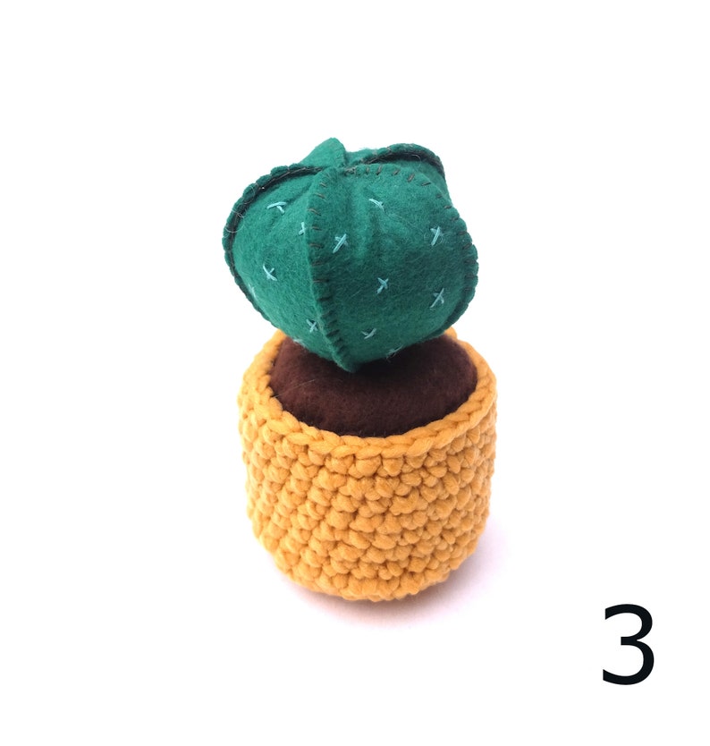 May include: A green felt cactus with brown felt soil in a yellow crocheted pot. The cactus has a stitched pattern of crosses.