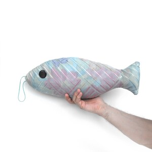 Peut inclure: Un coussin en forme de poisson ray&eacute; bleu et rose avec un &oelig;il en bouton noir. Le coussin est tenu par une main.