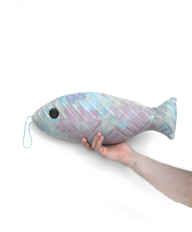 Peut inclure: Un coussin en forme de poisson ray&eacute; bleu et rose avec un &oelig;il en bouton noir. Le coussin est tenu par une main.