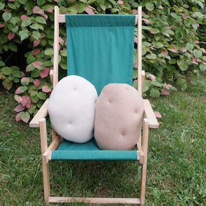May include: Two plush pillows shaped like potatoes, one white and one brown, are sitting on a green canvas chair with wooden arms. The chair is outdoors on a grassy surface.