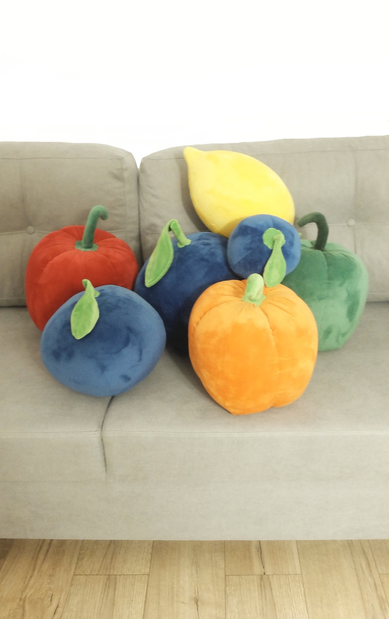 May include: A group of plush fruit-shaped pillows in various colors, including red, orange, yellow, green, and blue. The pillows are arranged on a light gray couch.