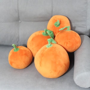 May include: Five plush orange pumpkins with green stems and leaves rest on a gray sofa. The pumpkins vary in size, creating a cozy, autumnal display. The soft, round shapes and vibrant colors evoke a harvest theme.