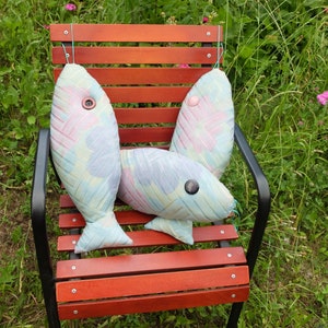 Peut inclure: Trois coussins en forme de poisson color&eacute;s avec des yeux en boutons sont pos&eacute;s sur un banc en bois avec un cadre en m&eacute;tal noir. Le banc est entour&eacute; d'herbe verte.