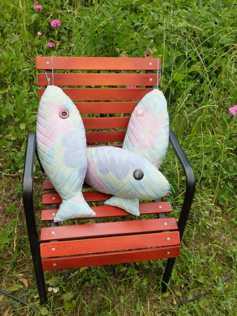 Peut inclure: Trois coussins en forme de poisson color&eacute;s avec des yeux en boutons sont pos&eacute;s sur un banc en bois avec un cadre en m&eacute;tal noir. Le banc est entour&eacute; d'herbe verte.