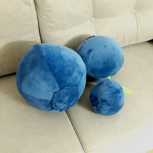 May include: Three plush blueberry pillows in various sizes, the largest is a deep blue, the medium is a lighter blue, and the smallest is a dark blue with a green stem. The pillows are sitting on a light beige couch.