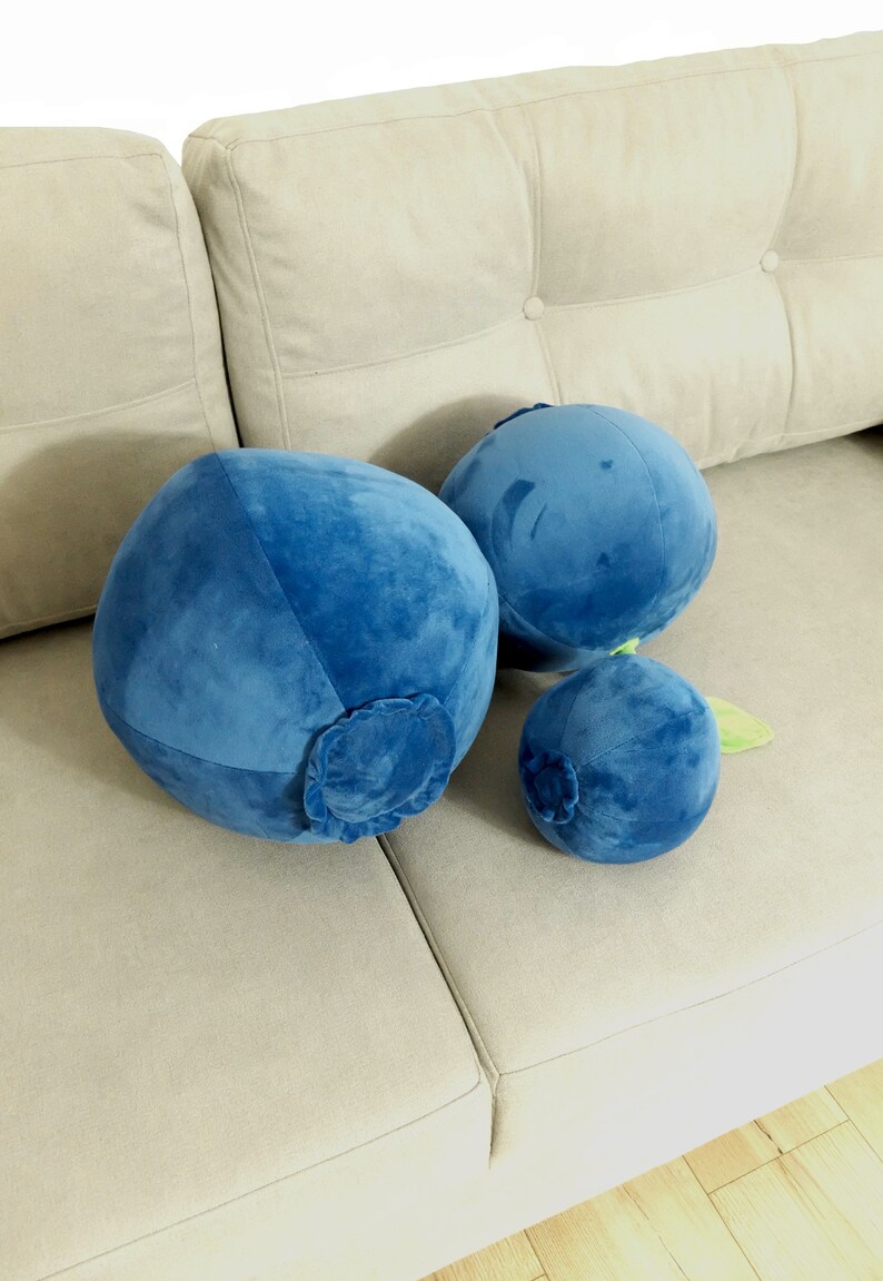 May include: Three plush blueberry pillows in various sizes, the largest is a deep blue, the medium is a lighter blue, and the smallest is a dark blue with a green stem. The pillows are sitting on a light beige couch.