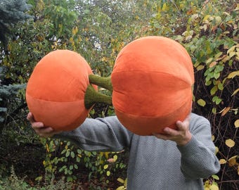 May include: A person holding two large orange plush pumpkins with green stems. The pumpkins are about the size of a person's head.